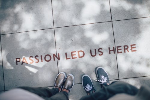 Picture of sidewalk with the words "passion led us here" written on it. Two people are standing below the words showing shoes and legs only.