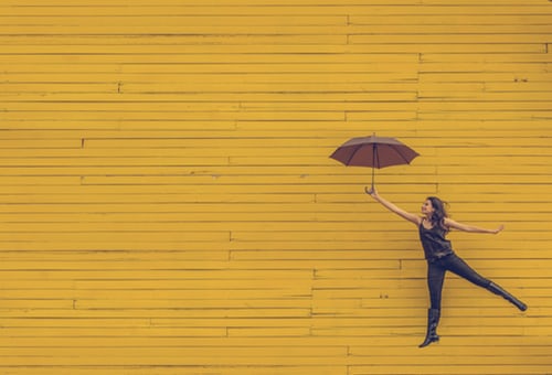 A yellow background with a woman holding an umbrella jumping the air to depict excitement on how to become an Excel Expert.