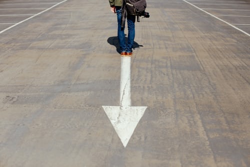 Photo of road with a white arrow and a man with a backpack adjacent to the arrow only depicting his waist down.