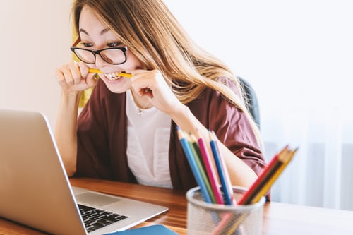 Woman at desk wearing glasses with a cup of pencils looking at a laptop with one pencil in her mouth.